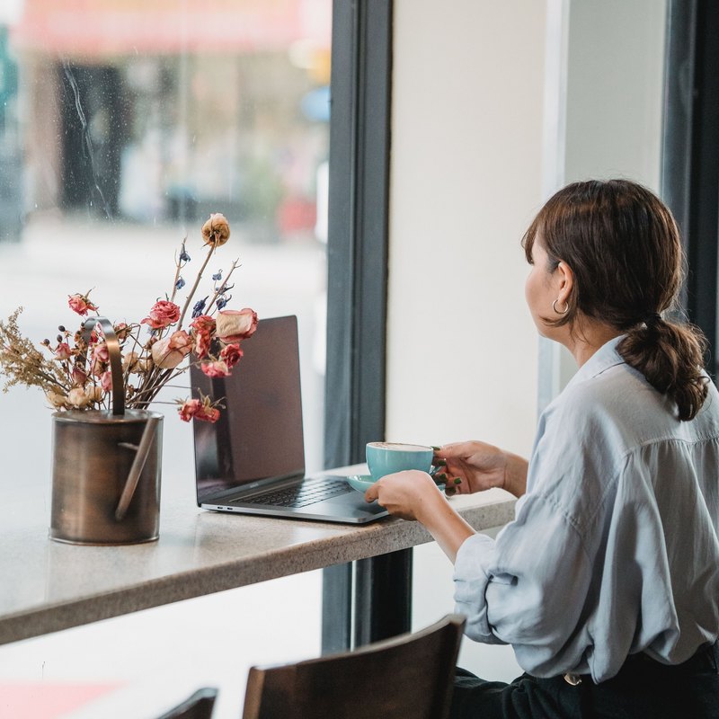Woman holding a turquoise cup beside a laptop at a cafe window