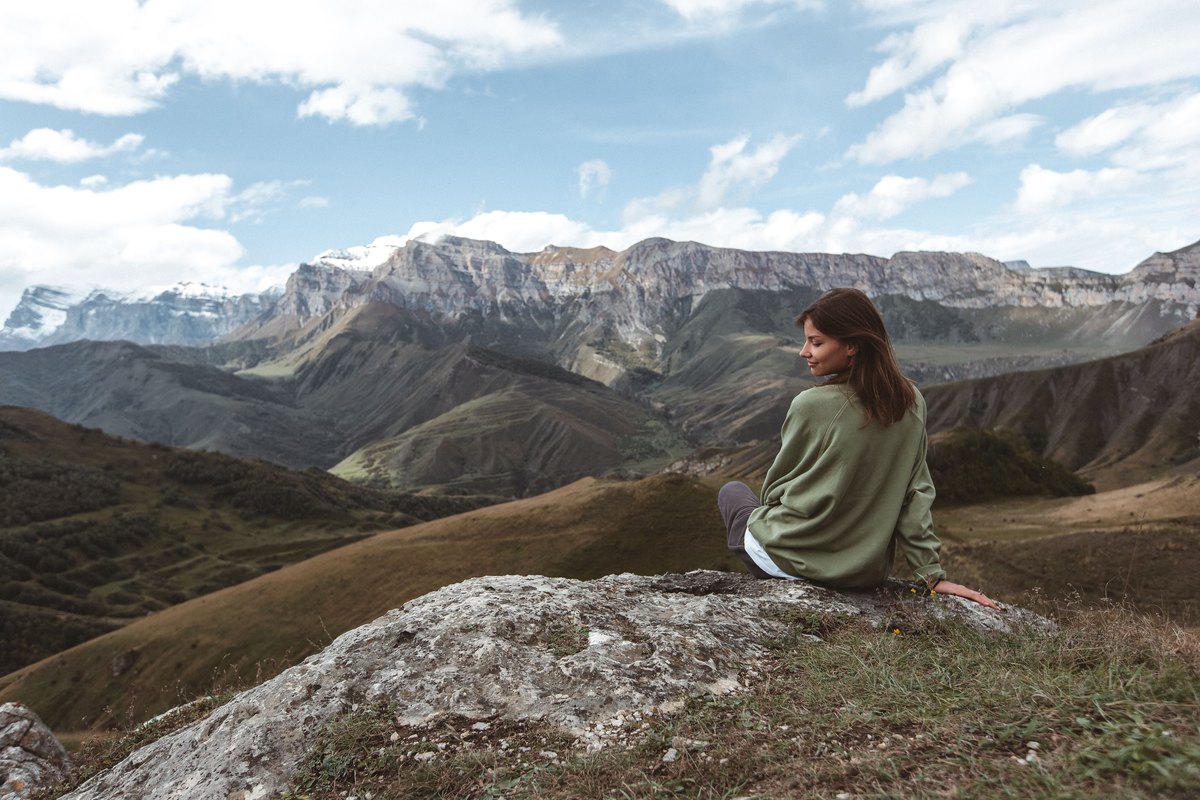 Woman in a green sweatshirt sitting on a rock above green valleys and a mountain ridge