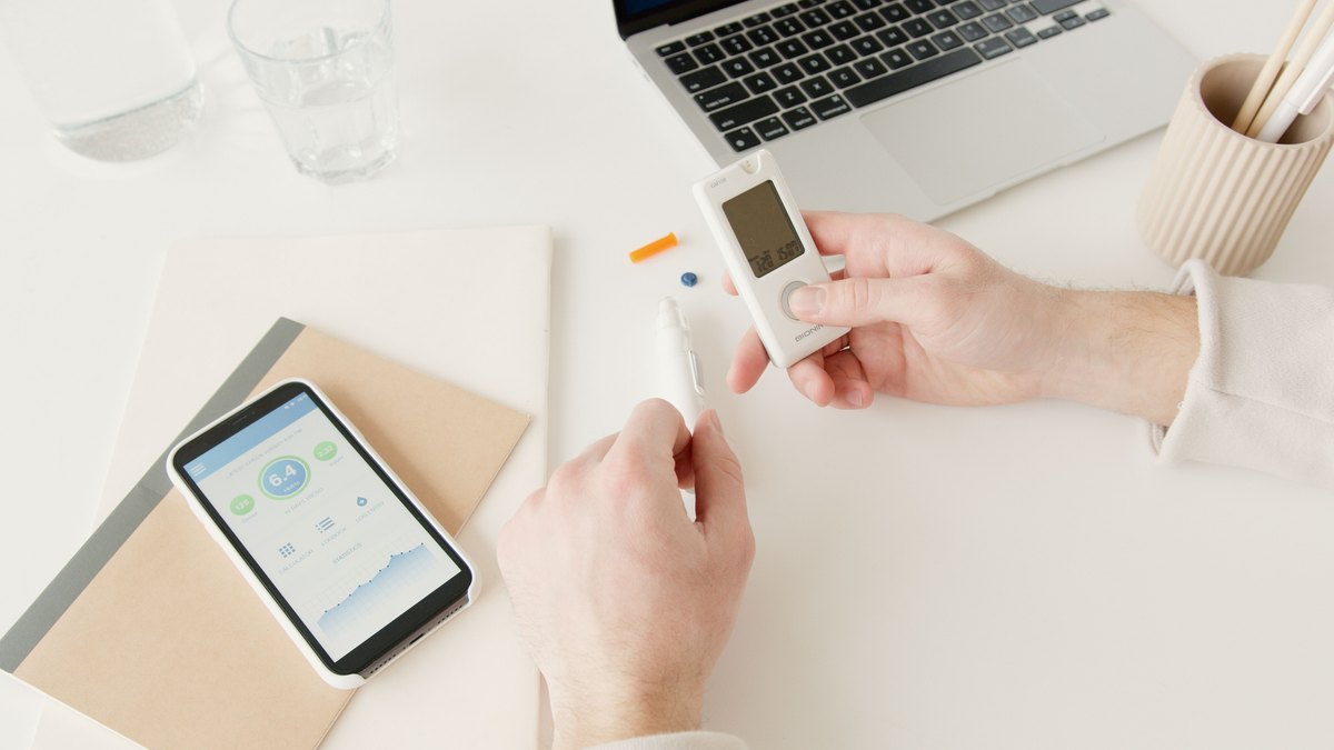 Hands using a glucose meter beside a laptop and phone at home