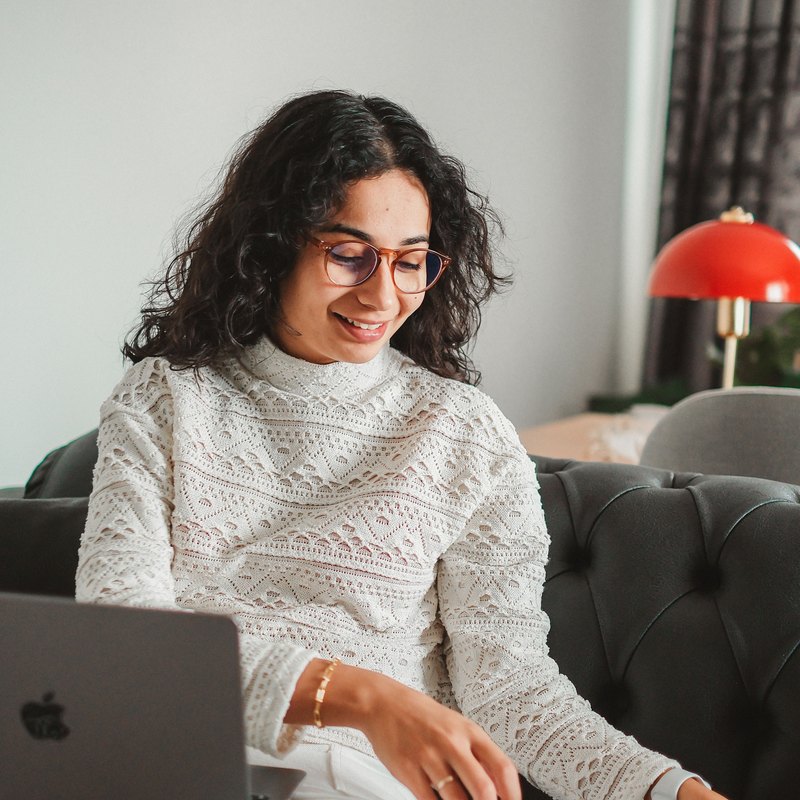 Smiling woman in glasses on a sofa with a laptop on her lap, reaching for an open book