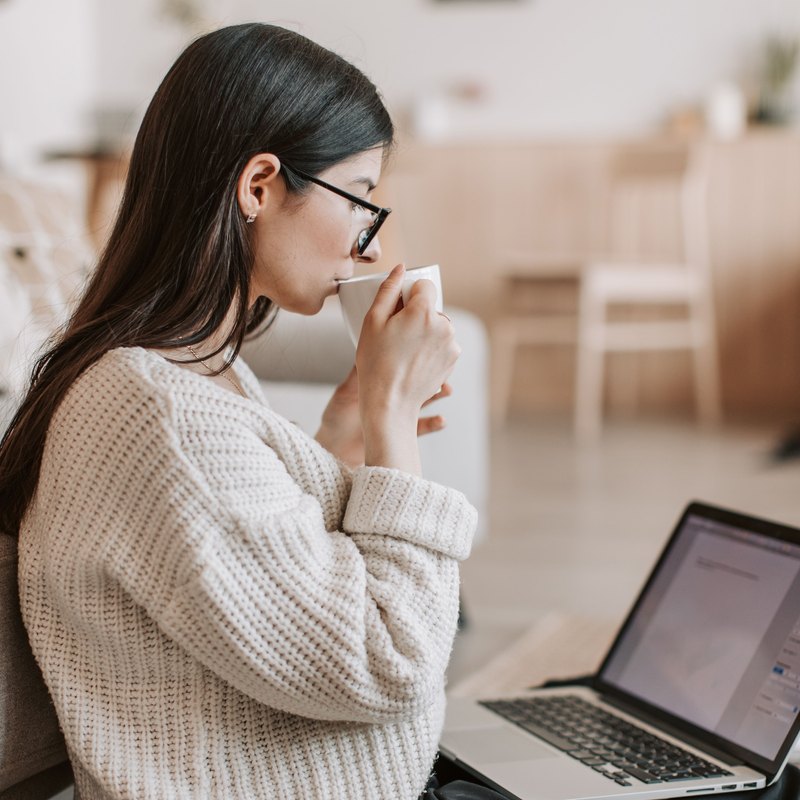 Woman in a cream sweater drinking tea with a laptop on her legs