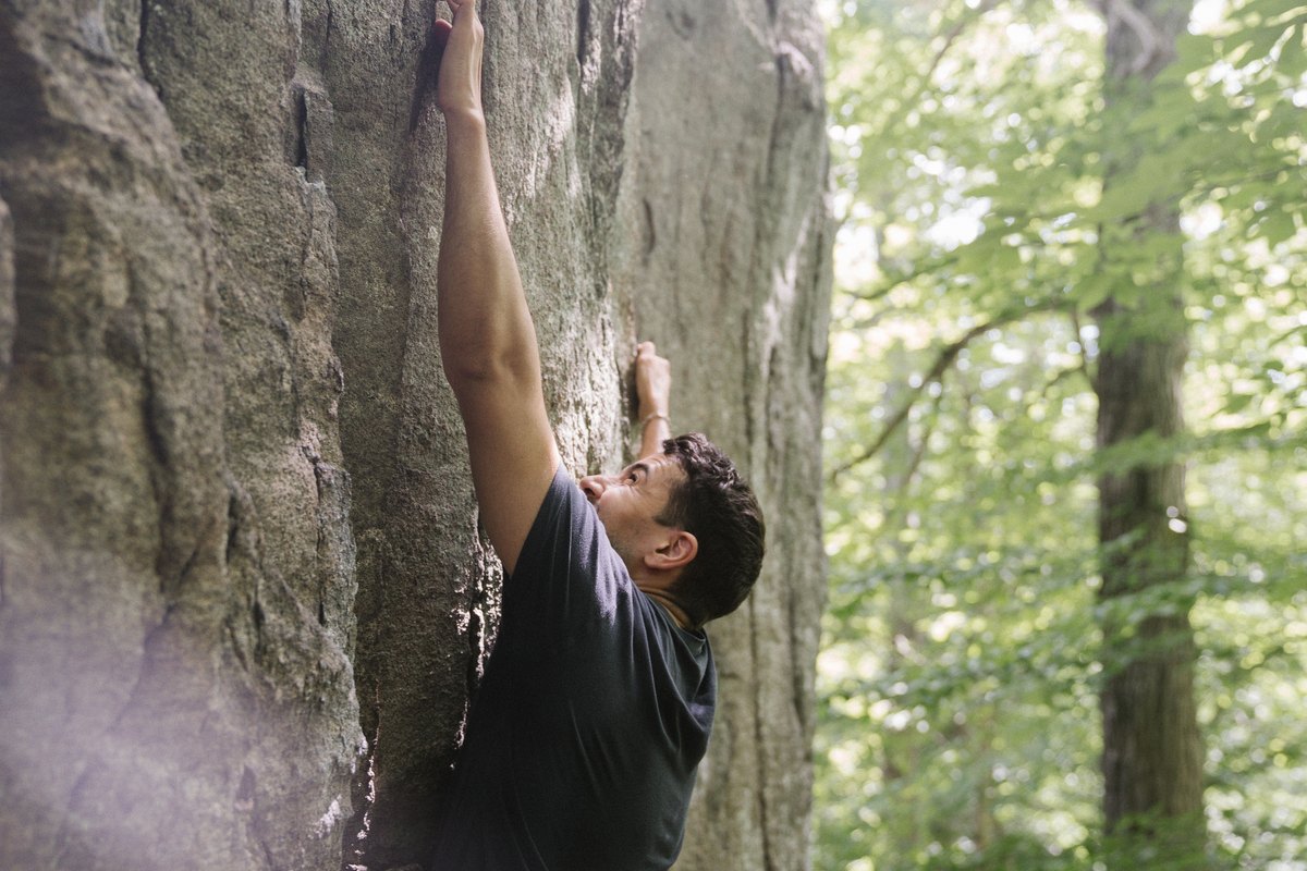 Man in a black T-shirt bouldering on a gray rock face in a forest