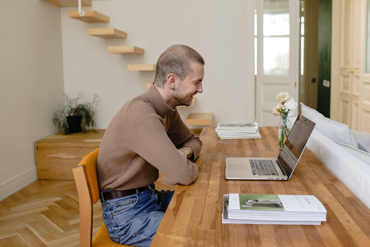 Smiling man in a brown sweater talking over a laptop at a wooden table